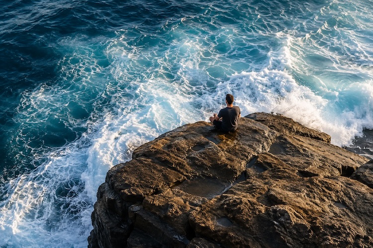 Person sitting steadily on coastal rocks as powerful ocean waves move around them